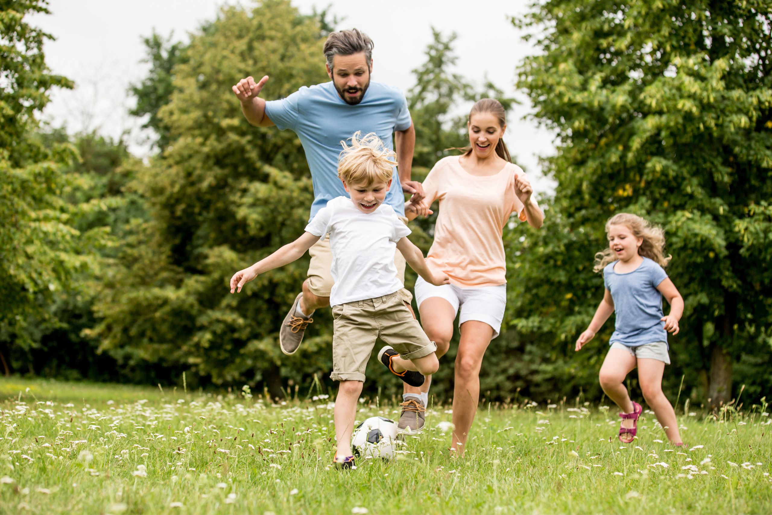 A mom, dad and their two children laughing and kicking a soccer ball around against a beautiful forest setting in a park